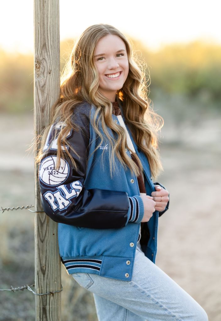 Senior portrait of girl in letterman jacket leaning against wood post with golden sun backlighting. Portrait by Lane and Mistie Photography in Midland TX.