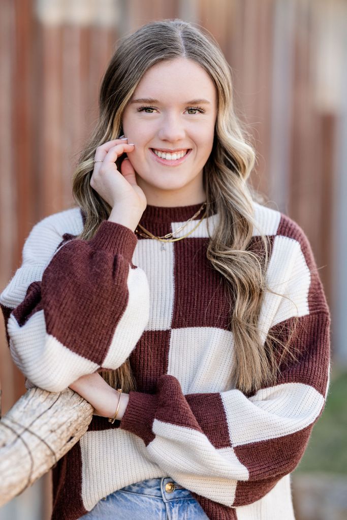 Senior portrait of girl in block sweater leaning on fence with hand on face pose. Portrait by Lane and Mistie Photography in Midland TX.